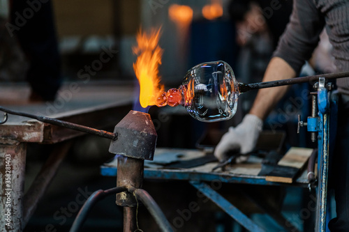 Close-up of a glassblower artisan shaping the hot molten glass at strong fire inside his workshop. Manual glass processing by the craftsmen inside a glass factory in Transylvania, Romania. 