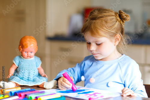 little alone toddler girl painting with felt pens during pandemic coronavirus quarantine disease. Happy creative child with old vintage doll, homeschooling and home daycare with parents