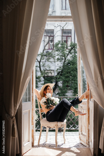 Stylish portrait of a girl on a vintage balcony who covers her breast with fresh peonies and on her head a stylish straw slag with a black ribbon around her face