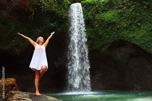 Young woman travel in Bali rainforest. Happy girl enjoy jungle nature. Stand in natural pool under waterfall  see on falling water. Walking day tour  hiking activity adventure on family summer holiday