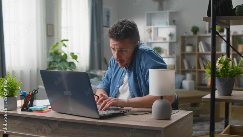 A young man speaks on a smartphone and uses a laptop while sitting at a desk in the living room A young man speaks on a smartphone and uses a laptop while sitting at a desk in the living room