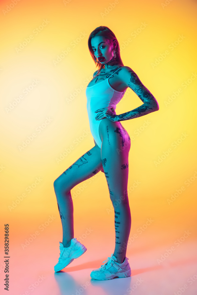 Beauty. Young caucasian woman in swimsuit posing sensual on gradient yellow background in neon. Beautiful model with tattoos. Human emotions, sales, ad concept. Resort and vacation, summertime.