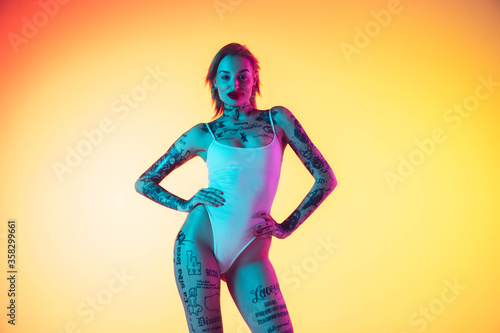 Confident. Young caucasian woman in swimsuit posing sensual on gradient yellow background in neon. Beautiful model with tattoos. Human emotions, sales, ad concept. Resort and vacation, summertime.