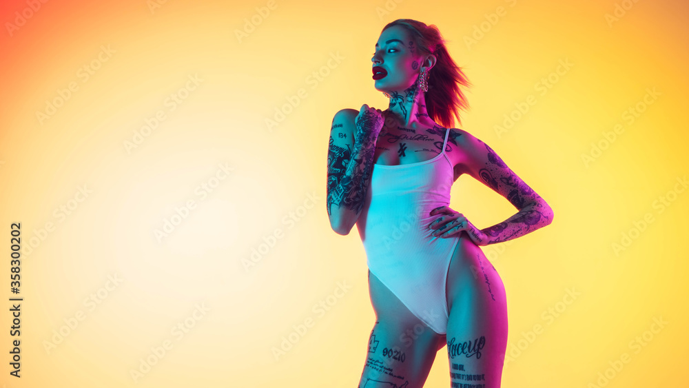 Young caucasian woman in swimsuit posing sensual on gradient yellow background in neon. Beautiful model with tattoos. Human emotions, sales, ad concept. Resort and vacation, summertime. Flyer.