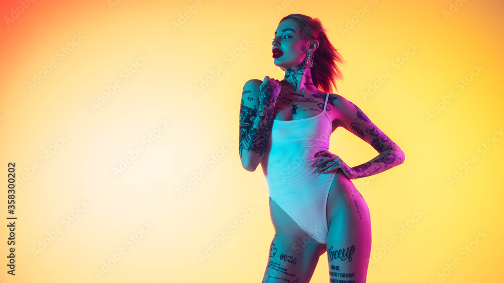 Young caucasian woman in swimsuit posing sensual on gradient yellow background in neon. Beautiful model with tattoos. Human emotions, sales, ad concept. Resort and vacation, summertime. Flyer. Young caucasian woman in swimsuit posing sensual on gradient yellow background in neon. Beautiful model with tattoos. Human emotions, sales, ad concept. Resort and vacation, summertime. Flyer.