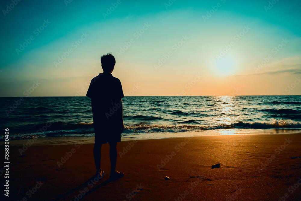 Young adult man standing on sand and staring at small waves of sea and orange sunset.