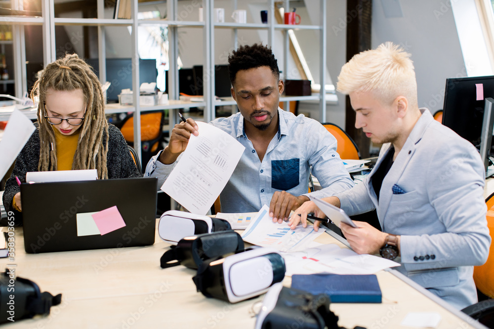 Office life, teamwork. Three young multiethnical business people, blond girl with dreadlocks, African and Caucasian men, sitting at the table, discussing something, working with project papers