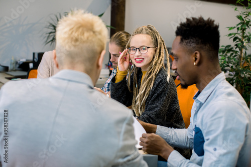 Team of hipster skilled multiethnic developers, students, working on business strategy, while sitting in modern office. Pretty smiling blond girl in eyeglasses and with long dreadlocks looks at camera