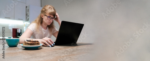 Young woman working remotely using a laptop. She has many tasks. She is focused and looks at the monitor, does not have time to drink coffee and eat a bun with poppy seeds. Copy space
