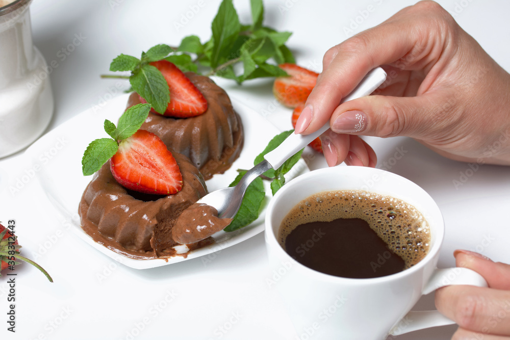 Woman eats dessert. Chocolate fudge garnished with half a strawberry with petals. Garnished with sprigs of mint and strawberries. Near a cup of coffee. On the white surface of the table. Woman eats dessert. Chocolate fudge garnished with half a strawberry with petals. Garnished with sprigs of mint and strawberries. Near a cup of coffee. On the white surface of the table.