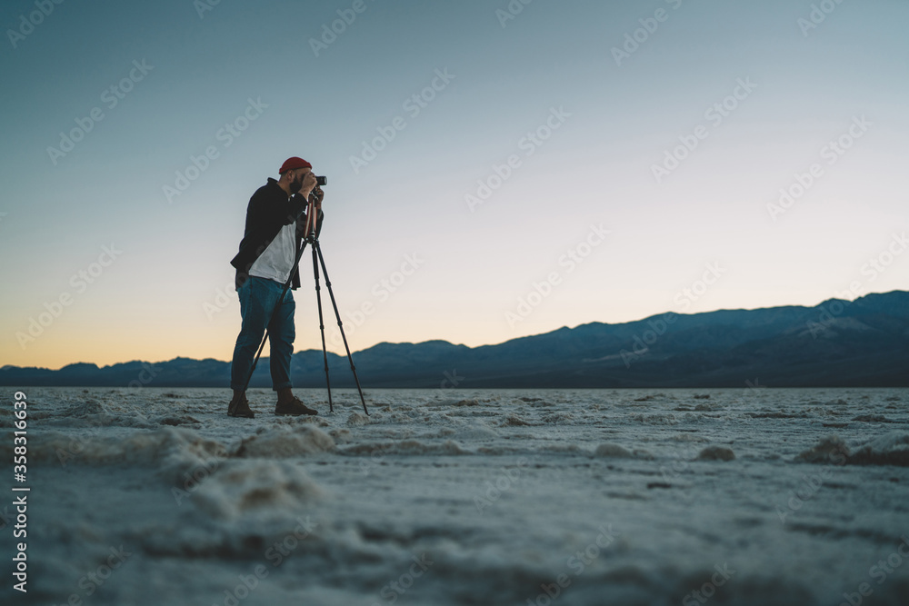 Skilled male journalist taking picture in expedition in death valley explore wild lands and environment, professional man photographer taking picture on equipment with tripod on sundown in desert