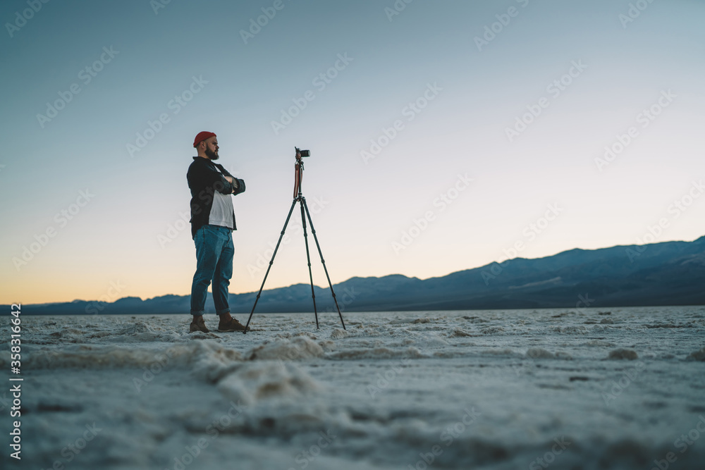 Concentrated male photographer standing near tripod shooting breathtaking landscape of Badwater basin on sunset, serious hipster guy making photo on modern camera during expedition in death valley