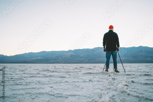 Back view of man using tripod for making pictures of breathtaking view of death valley landscape, hipster guy looking at high rocks on horizon at evening dusk having expedition in Badwater basin.