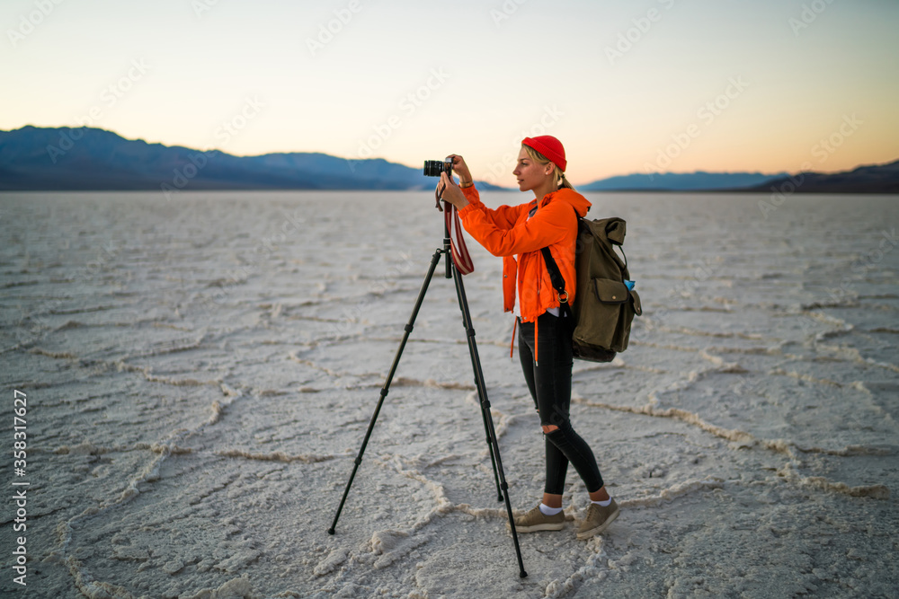Hipster girl wanderlust enjoying scenery of evening desert lands using tripod for shooting video of sunset, skilled female photographer making pictures of wild nature and landscapes during trip