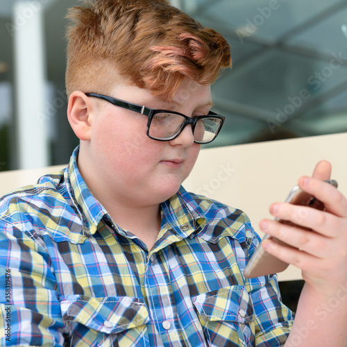 Red-haired little boy in a plaid shirt with smartphone sitting at a bar table. Cute red-haired boy with cellphone and glasses in an urban context.