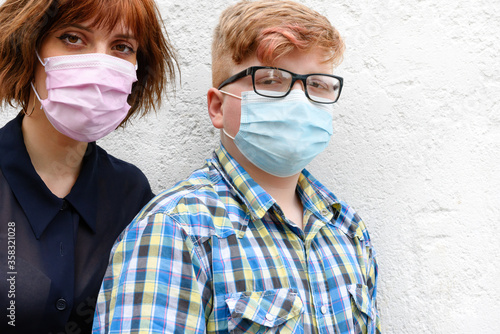 Young red-haired woman and boy wearing a surgical mask stand against a wall background.