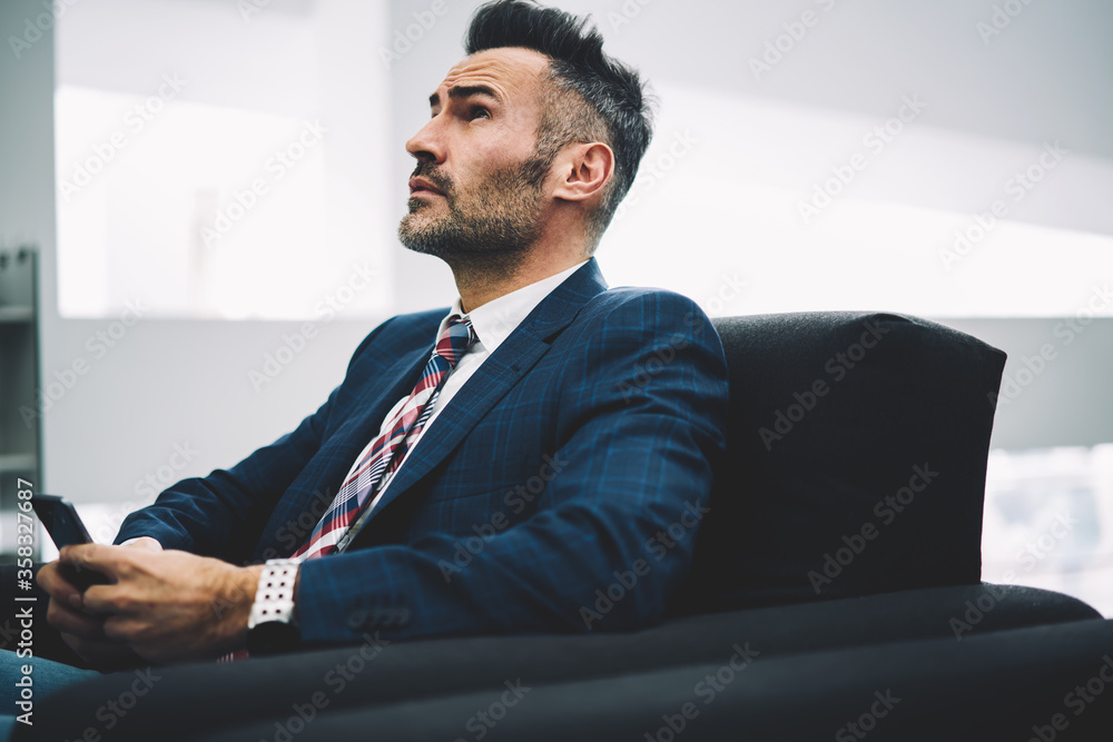 Thoughtful proud ceo looking up while thinking on text answer in message sitting on couch with smartphone in hands.Pondering mature businessman in formal wear chatting on cellular in office Thoughtful proud ceo looking up while thinking on text answer in message sitting on couch with smartphone in hands.Pondering mature businessman in formal wear chatting on cellular in office