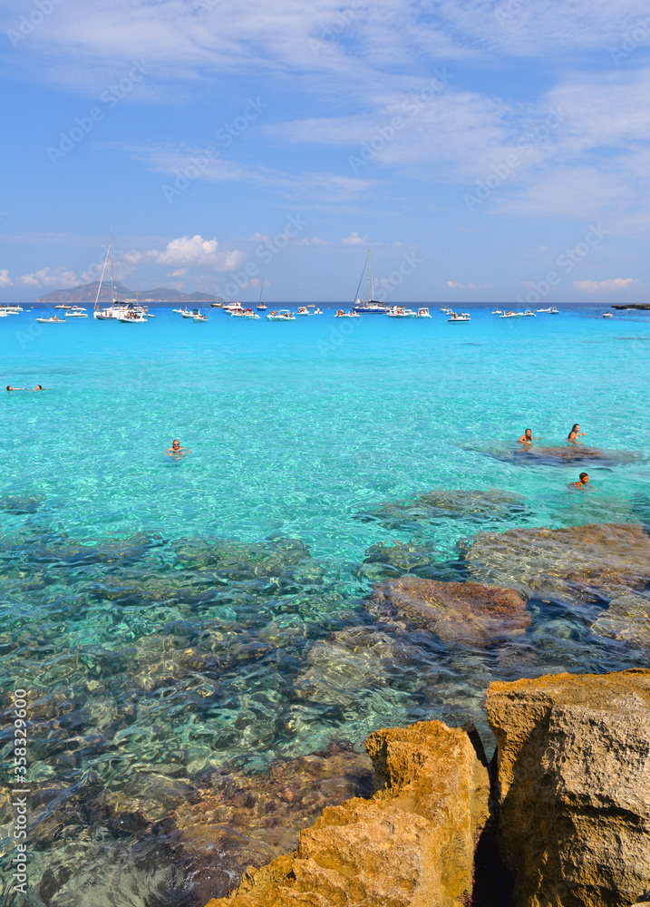 paradise clear torquoise blue water with boats and cloudy blue sky in background in Favignana island, Cala Rossa Beach, Sicily South Italy.