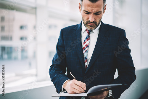 Successful proud ceo middle aged making notes in notepad standing in office interior.Confident experienced mature financial director dressed in formal wear signing accounting documents