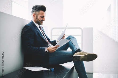 Talented experienced male chief editor of popular magazine reading text of analytic article about international relations and business affairs admiring work of skilled author sitting in office