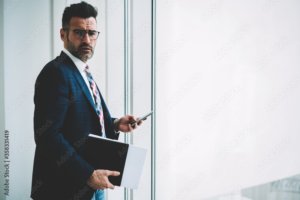 Half length portrait of confident mature entrepreneur dressed in formal wear checking mail on smartphone device connected to 4G internet standing near publicity area for your business information Half length portrait of confident mature entrepreneur dressed in formal wear checking mail on smartphone device connected to 4G internet standing near publicity area for your business information