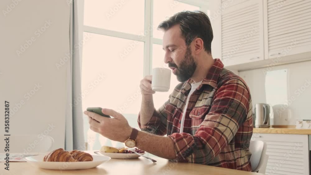 Handsome young positive optimistic bearded man at the kitchen indoors at home having a breakfast while using cellphone Handsome young positive optimistic bearded man at the kitchen indoors at home having a breakfast while using cellphone