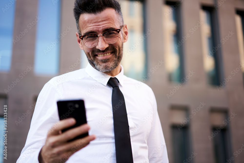 Selective focus, happy 40 years old manager dressed in fashionable white shirt searching financial news and sending message using smartphone and high speed internet connection near trade company Selective focus, happy 40 years old manager dressed in fashionable white shirt searching financial news and sending message using smartphone and high speed internet connection near trade company