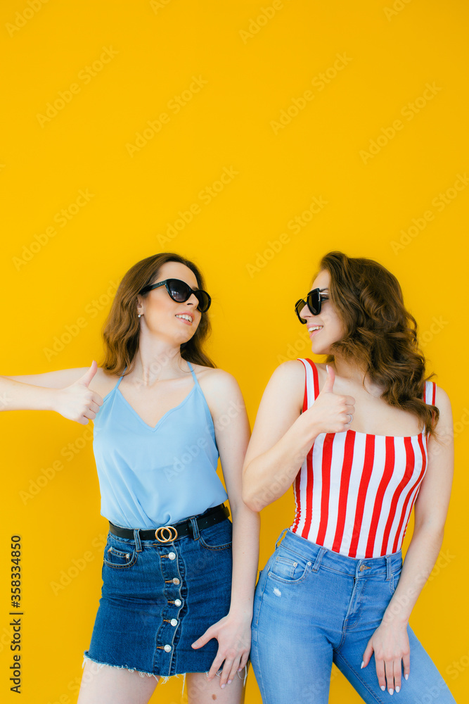 Two young beautiful blond smiling hipster girls in trendy summer clothes. Sexy carefree women posing near yellow wall in sunglasses. Positive models going crazy