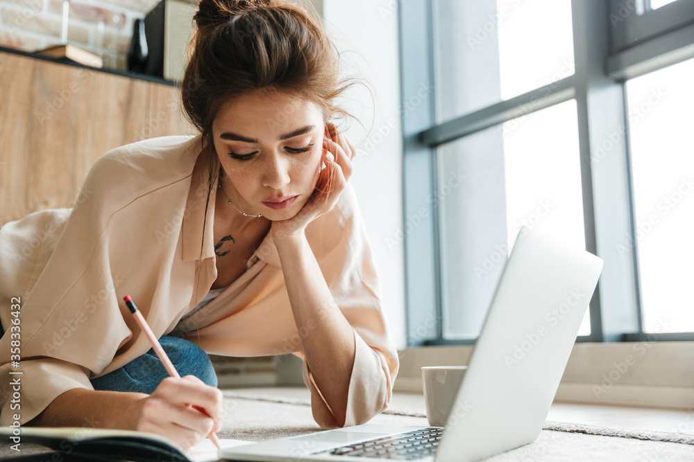 Obraz premium Image of woman writing down notes and using laptop while sitting on floor
