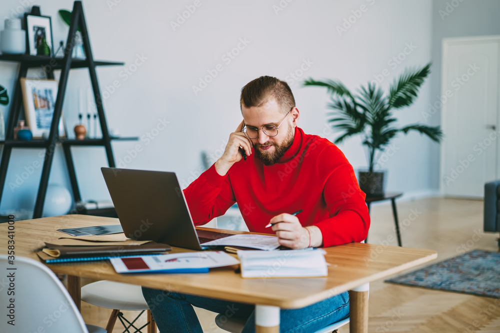 Obraz premium Young hipster guy making notes during telephone consultancy sitting at desktop,male freelancer making telephone call to banking operator for checking balance and planning project at home interior.