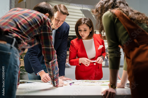 Team Of Architects Working On Project Paper Blueprint. Group Of Modern Business People In Casual Wear Discussing Architectural Designs While Sitting In The Creative Office.