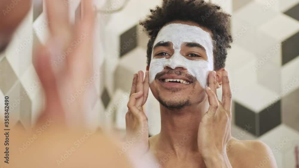 Young cheerful african guy taking care of his skin with face mask near mirror indoors in a bathroom Young cheerful african guy taking care of his skin with face mask near mirror indoors in a bathroom