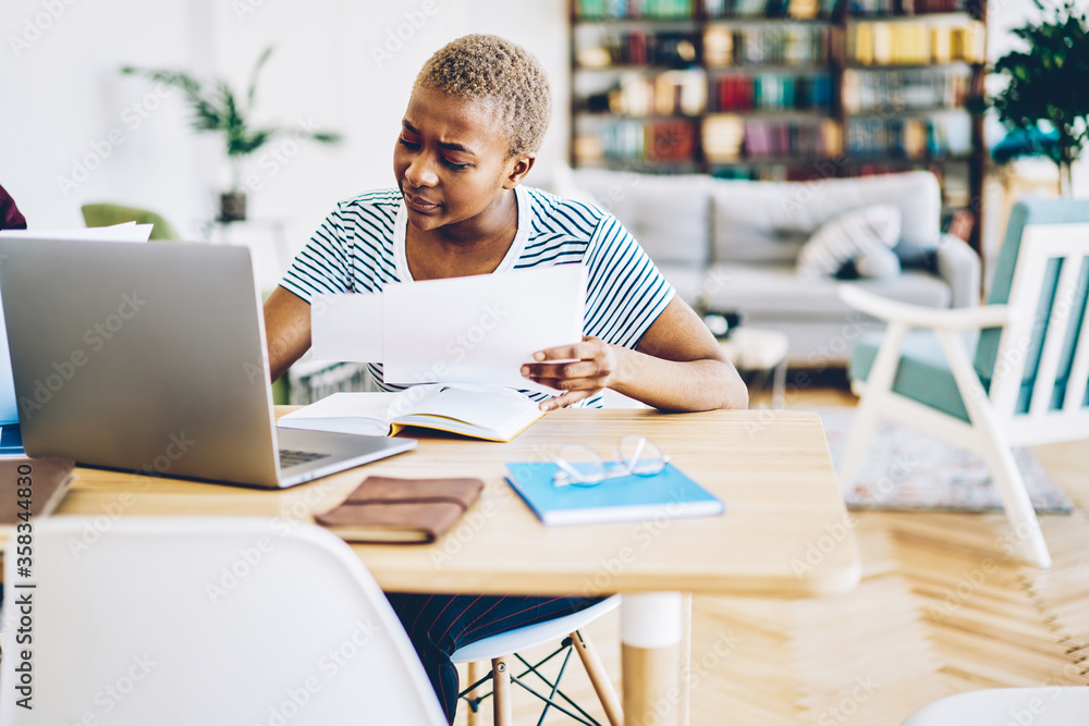 Obraz premium Concentrated african american woman making banking online using papers w with information, serious skilled dark skinned female freelancer doing remote job on laptop computer at home interior