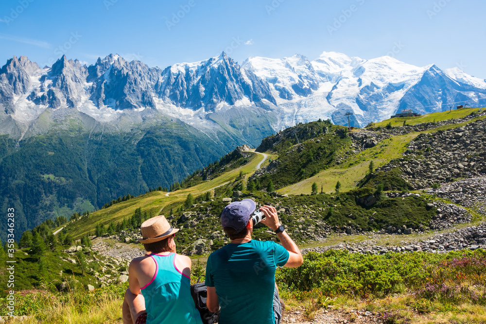 French  Alps summer travel. Couple of hikers (unidentified people, back view) admire beautiful snow covered Mont Blanc mountains range, resting before keeping to walk. France nature tourism background