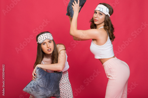 portrait of two excited girls dressed in pajamas having pillow fight isolated over pink background