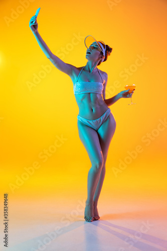 Fashion portrait of seductive girl in stylish swimwear on yellow background in neon. Summertime, beach season. Woman with cocktail taking selfie. Resort, vacation, holidays concept. Copyspace for ad.