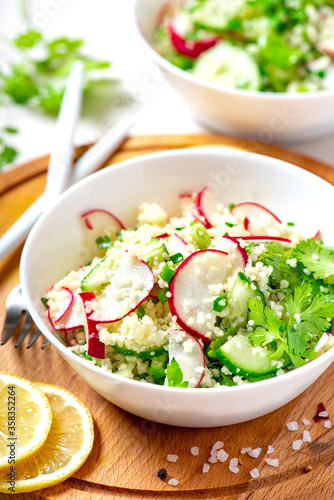 Fresh summer vegetable salad of couscous, radish, cucumbers, parsley and lemon in a white bowl on a served table. Vegan food. Spring or summer food. Salad with couscous and raw vegetables close-up.