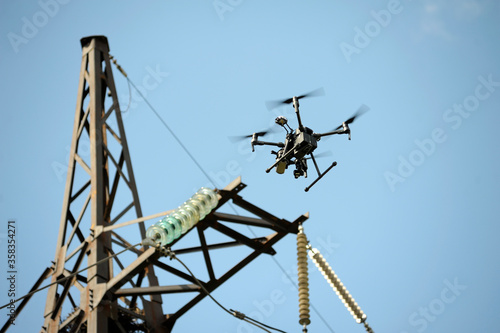 Pro quadcopter with camera aboard flying in a sky, electric pylon, cables and insulators on the background