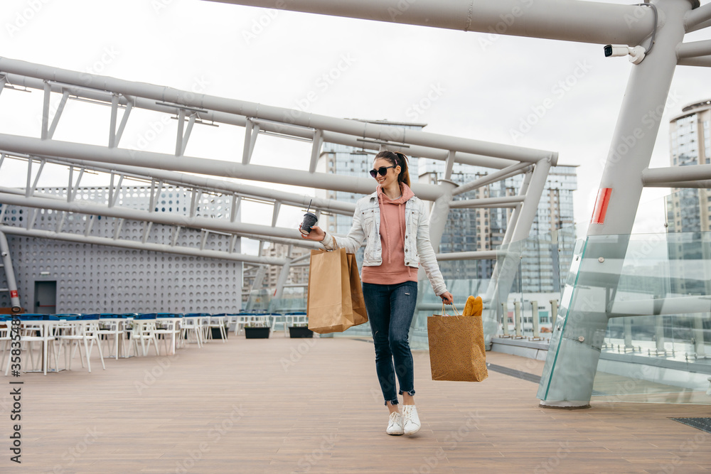 Smiling young woman holding coffee cup, walking with grocery shopping paper bag with long white bread baguette over city background