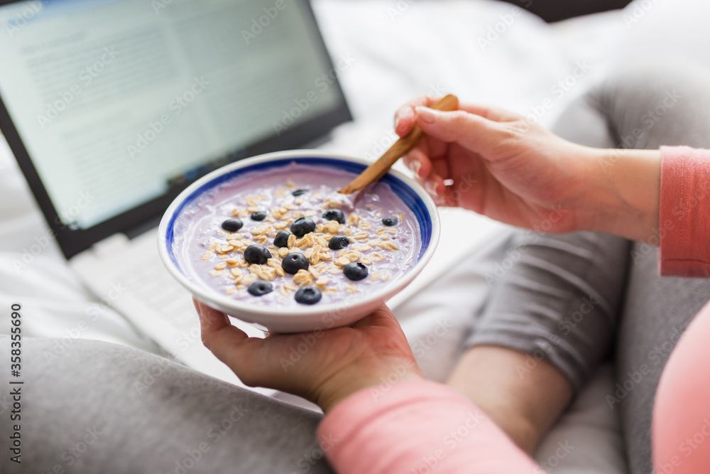 Close up of pregnant woman eating muesli with blueberries while working on laptop.