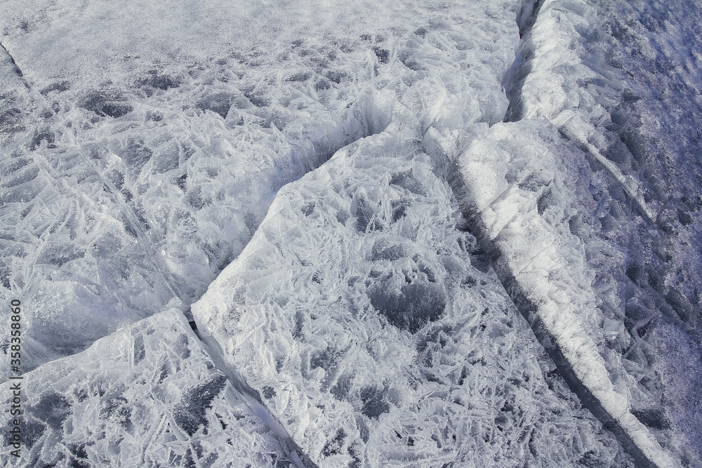 Naklejka premium Danger on the show surface: cracks in blue ice of cracked glacier in textured white snow surface under sunlight, closeup view, winter
