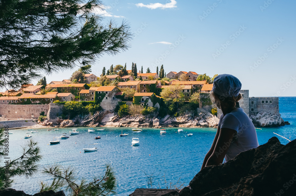 A girl in a white polka-dot panama admires the view of the island of St. Stephen in Montenegro. Sunny view of island among green thickets is surrounded by a blue lagoon with snow-white small boats.
