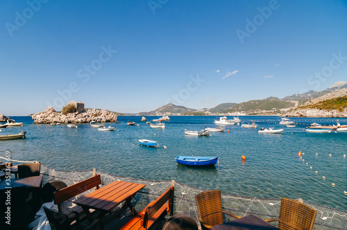 View of the lagoon in the Adriatic Sea from the beach cafe. In the foreground are wooden cafe tables and a fencing fishing net. In the background are low green hills, fishing boats in the sea.