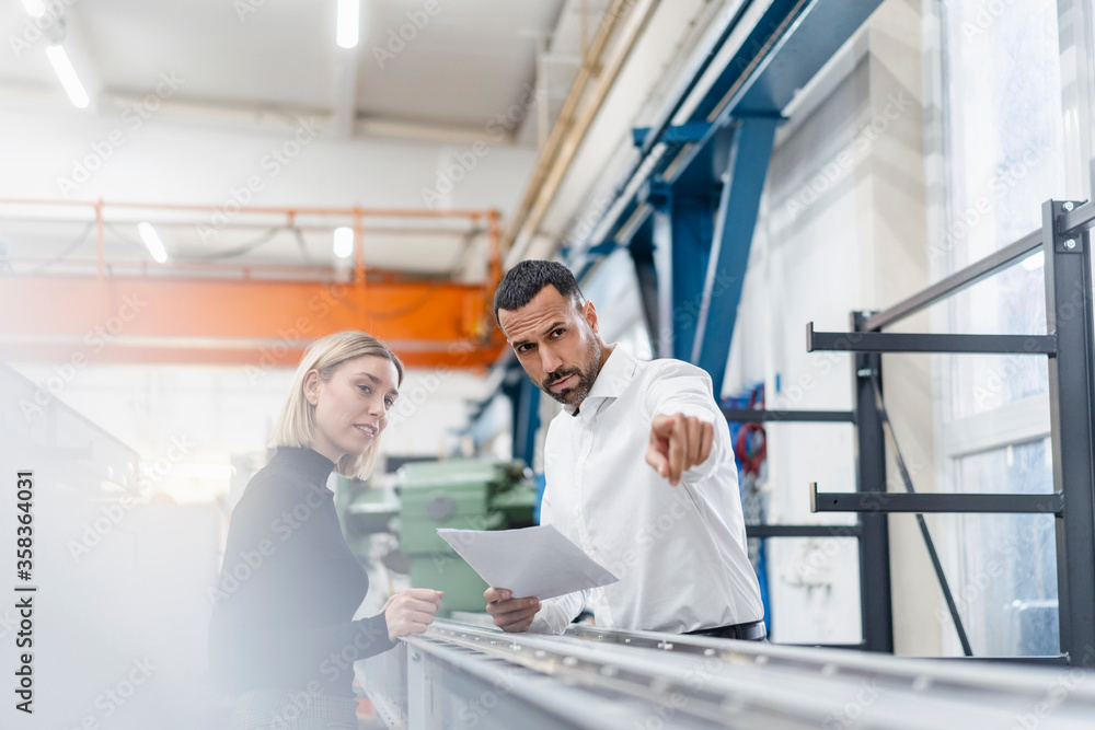 Businessman and woman with papers examining metal rods in factory hall Businessman and woman with papers examining metal rods in factory hall