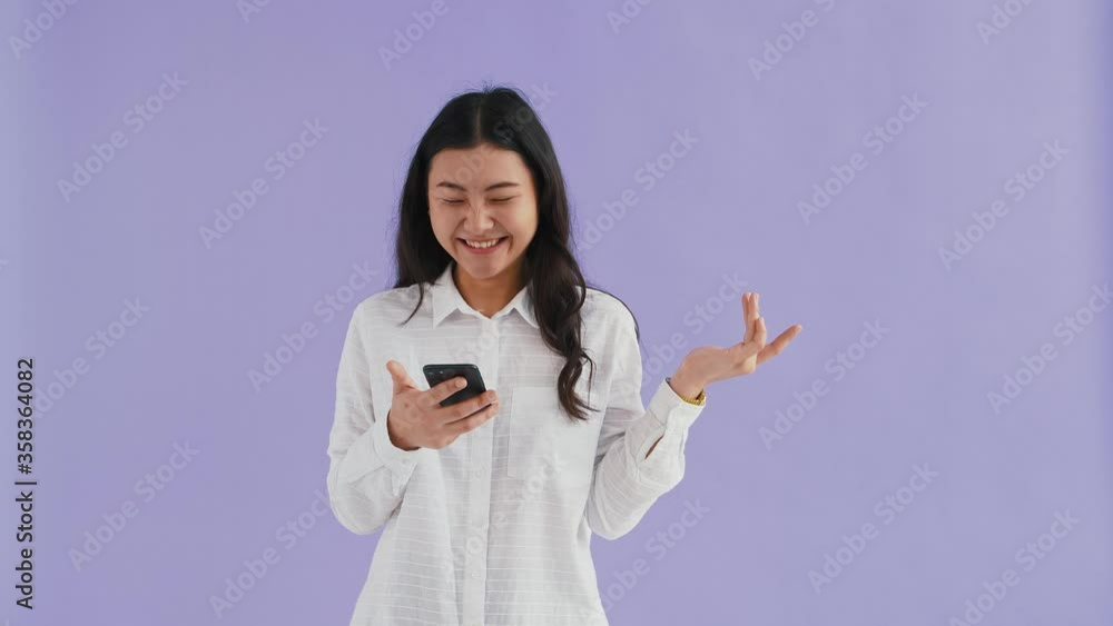 Asian female in white shirt. She using smartphone, smiling and looking surprised, saying wow. Posing against purple background. Close up, copy space Asian female in white shirt. She using smartphone, smiling and looking surprised, saying wow. Posing against purple background. Close up, copy space
