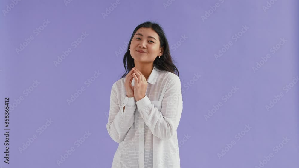 Gorgeous asian woman in white shirt. She smiling and playing her fingers like waiting for something pleasant. Posing on purple background. Close up Gorgeous asian woman in white shirt. She smiling and playing her fingers like waiting for something pleasant. Posing on purple background. Close up