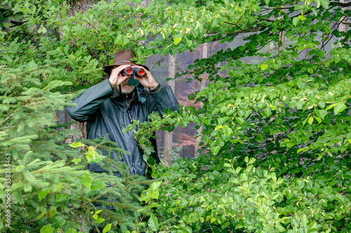 Tableau sur toile Well camouflaged between the bushes, a hunter observes the hunting area through his binoculars on a rainy day