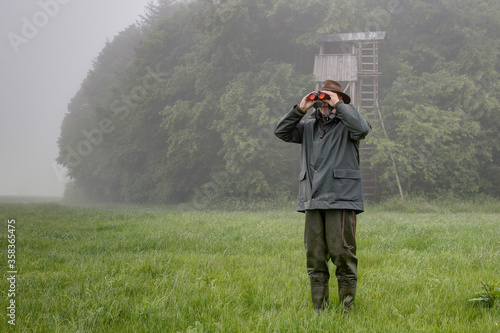 Foto On a rainy day a hunter stands in front of the hunting pulpit in the meadow and looks through his binoculars
