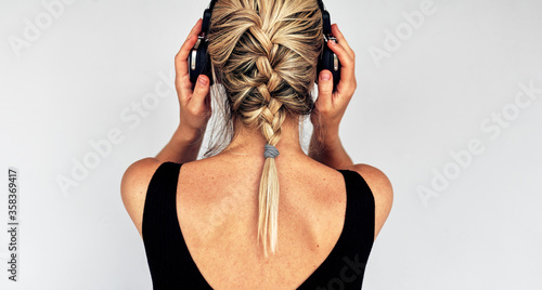 Rear view image of a blonde young woman touching her headphones during listening to music. Horizontal back view of pretty female in black dress with braid hairstyle listens to audiobook in headphones.