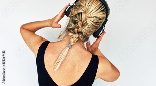 Studio rear view shot of attractive blonde young woman dancing and touching her headphones during listening to music. Beautiful female in black dress with braid hair listens to audiobook in headphones